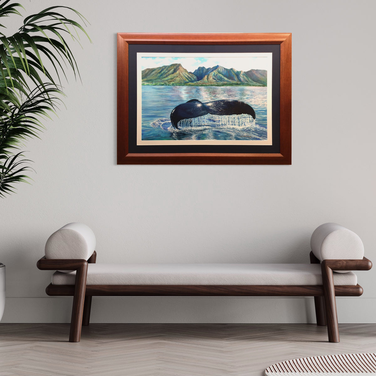 Professionally framed original watercolor art  depicting a humpback whale tail surfacing in Hawaiian blue water with green mountains in the background, hung on a light gray wall above a wooden bench with white cushions.
