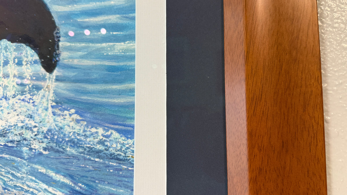Close-up of a framed and matted artwork showing blue ocean waves and part of a dark whale, alongside vertical color/material samples including a dark navy blue matting and white matting and a warm brown wood grain frame.