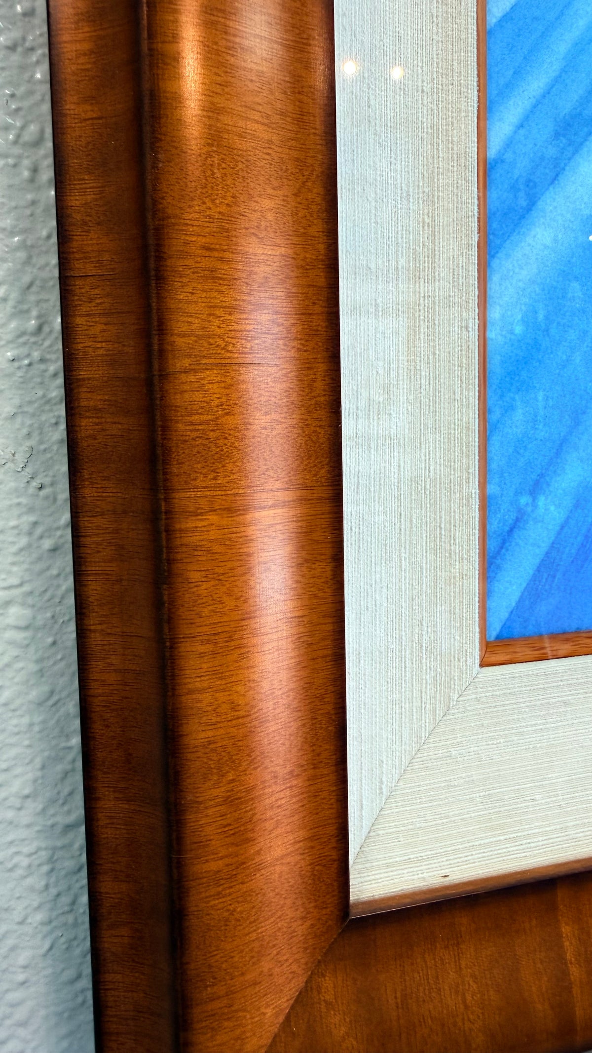 Close-up of a framed artwork corner showing a warm medium-brown Koa wood frame, a light natural textured silk inner fliner and small koa inner frame, and part of the blue ocean painted .