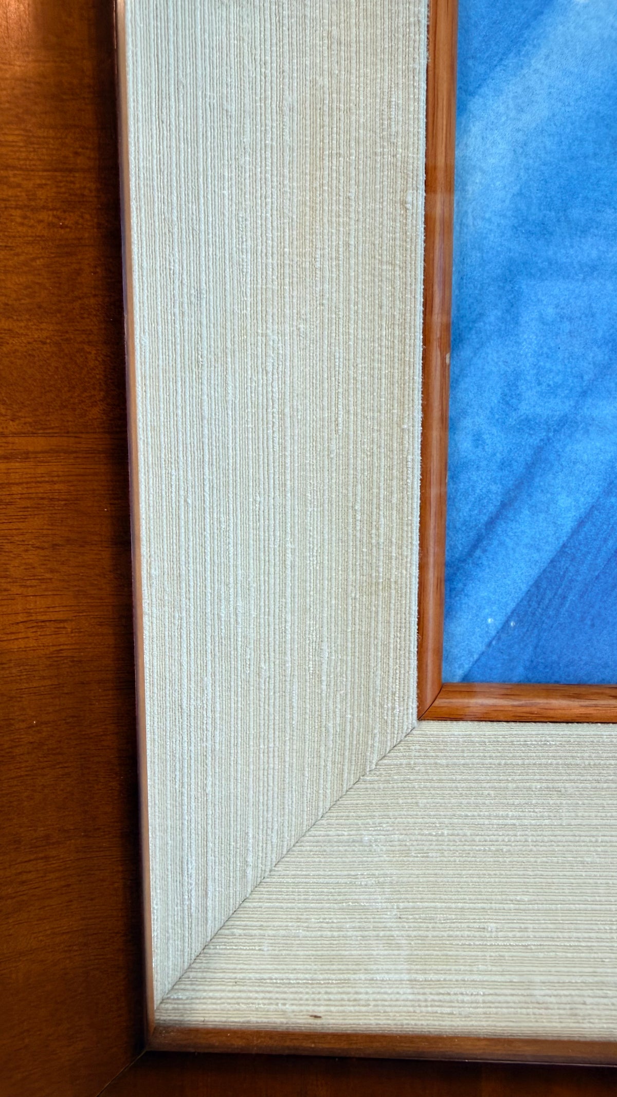 Close-up of a framed artwork corner showing a warm medium-brown Koa wood frame, a light natural textured silk inner fliner and small koa inner frame, and part of the blue ocean painted .