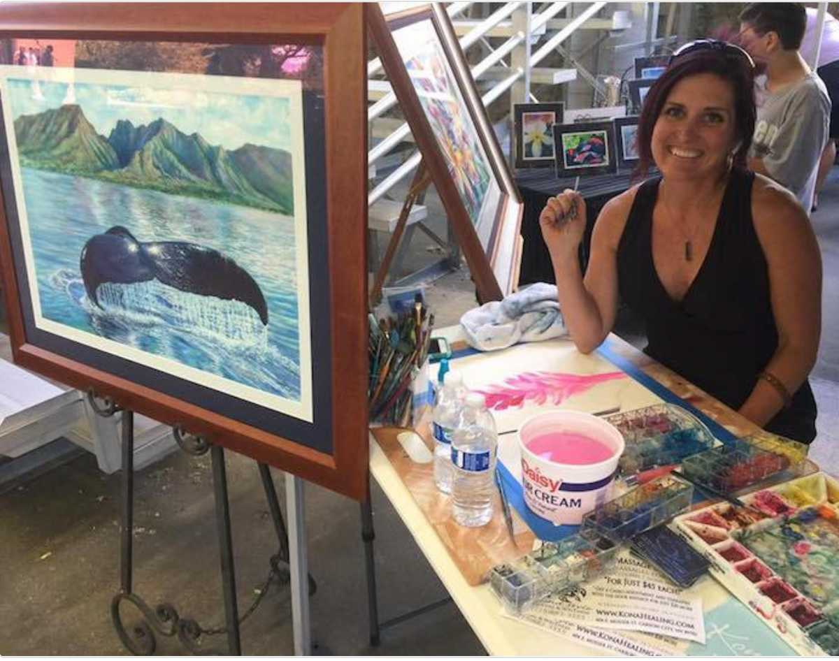 Christie Marie Elder with Sanctuary original watercolor smiling at an art show next to a large framed painting of a whale and mountains, with paint supplies and another easel in the background.